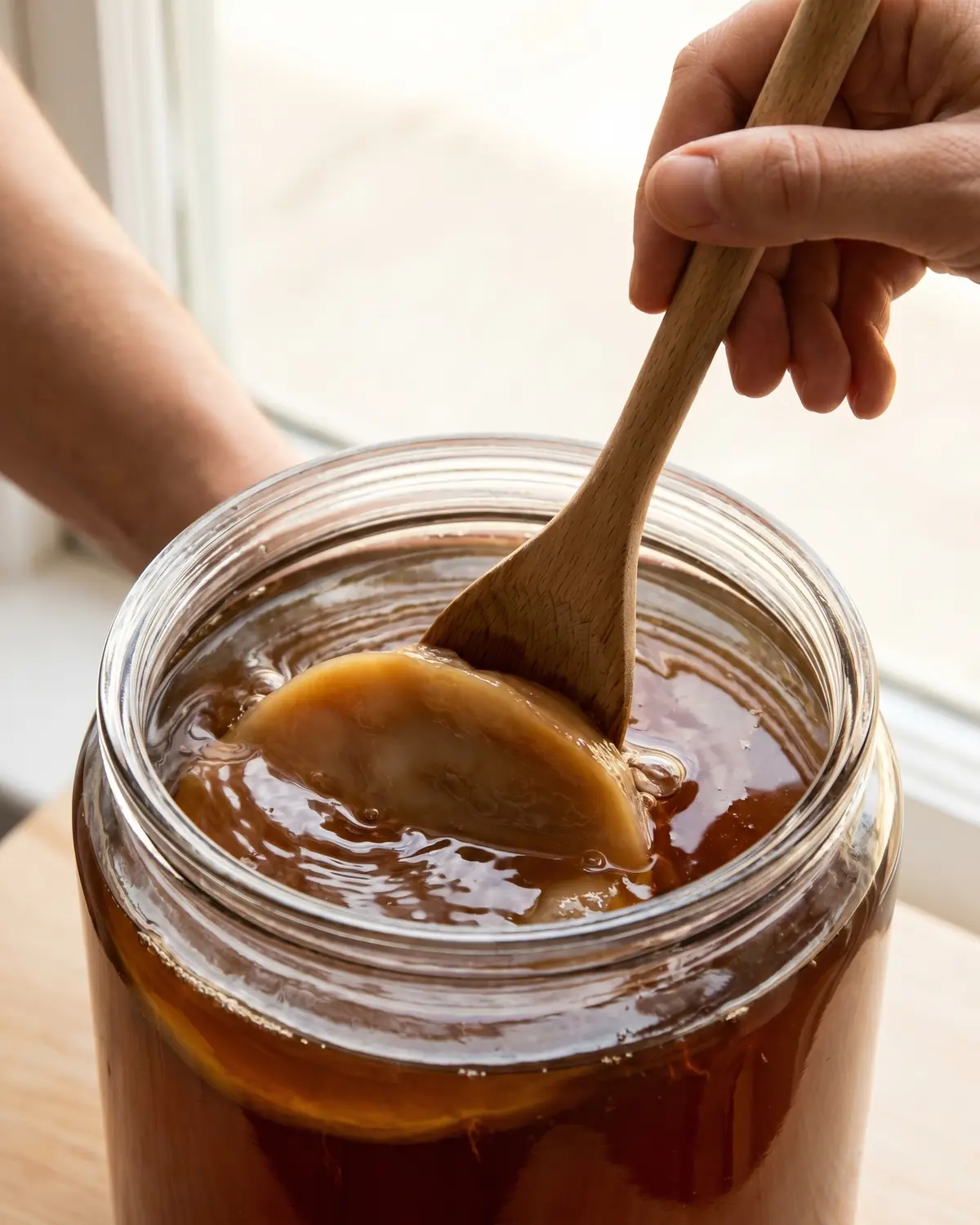 kombucha lidl Cucchiaio di legno che spinge delicatamente lo SCOBY sotto il tè ambrato durante la preparazione