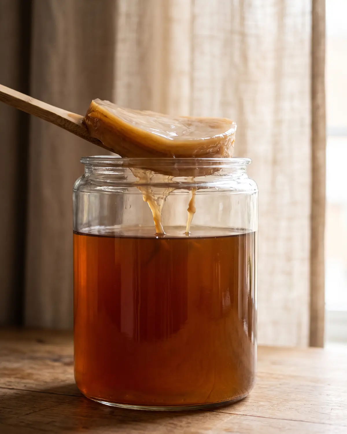 scoby hotel Barattolo di kombucha con SCOBY e filamenti morbidi, visto accanto a una tenda di lino sfocata.