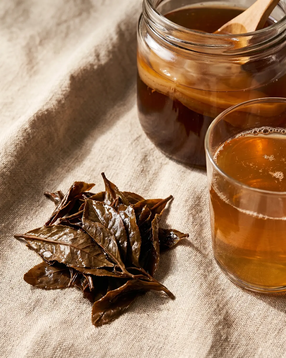 Foglie di t&egrave; umide accanto a un bicchiere di kombucha su una superficie di lino naturale.
