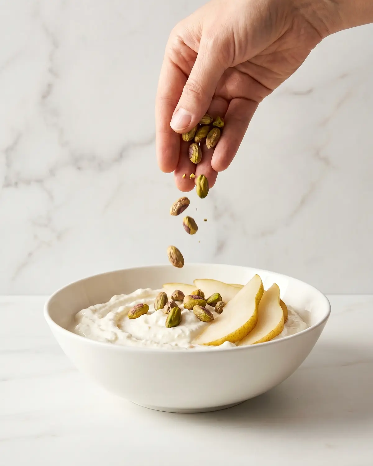yogurt con fermenti lattici Mano che aggiunge pistacchi su yogurt con pera, in una ciotola su marmo bianco in formato verticale.